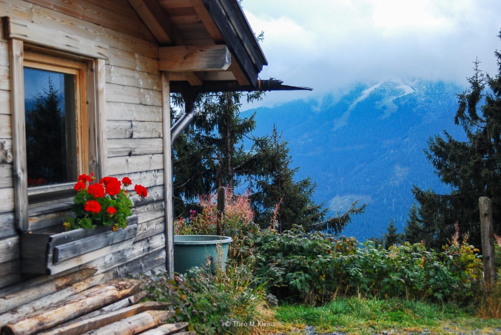 Almhütte mit roten Blumen am Fenster und Blick auf die umliegenden Berge